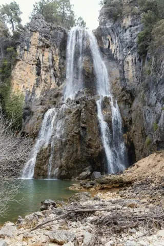 A stunning waterfall cascades down rugged cliffs into a clear pool, surrounded by rocky terrain and lush greenery in the Troodos Mountains, Cyprus.