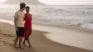 A couple walking barefoot along a sandy beach, with ocean waves in the background. The woman is wearing a red dress, and the man is dressed in a beige t-shirt and black shorts.