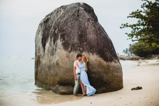A couple embracing near a large rock on a sandy beach, with ocean waves and palm trees in the background. The woman is wearing a flowing blue dress, and the man is dressed casually in jeans and a white shirt.