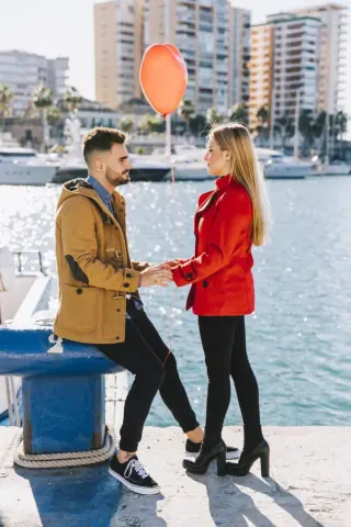 A couple standing by a marina holding hands, with the woman holding a red balloon. Luxury yachts and modern buildings are visible in the background.